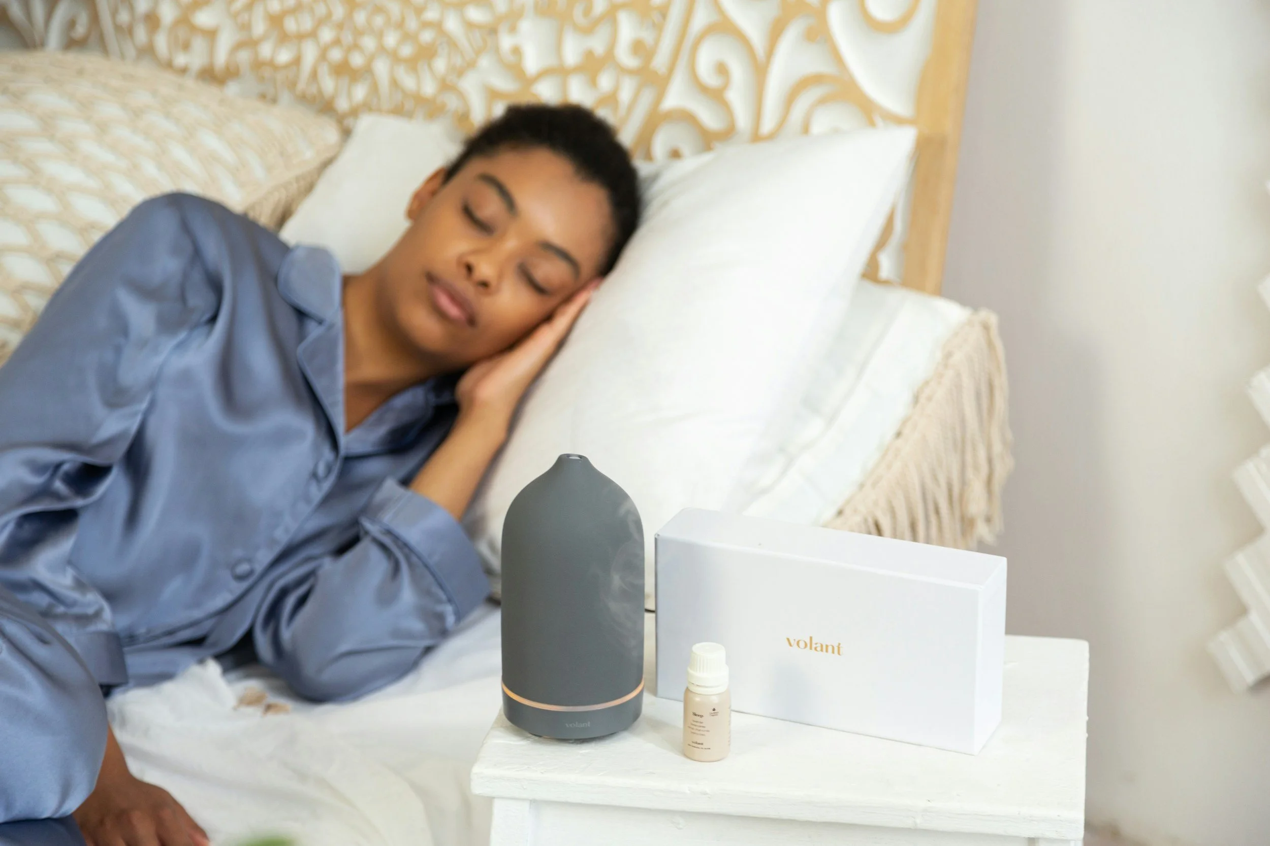 A woman of color with short dark hair, wearing a gray satin pajama top, sleeping on a white pillow in bed with a beige patterned headboard. In front of her on a small white table are small product bottles.
