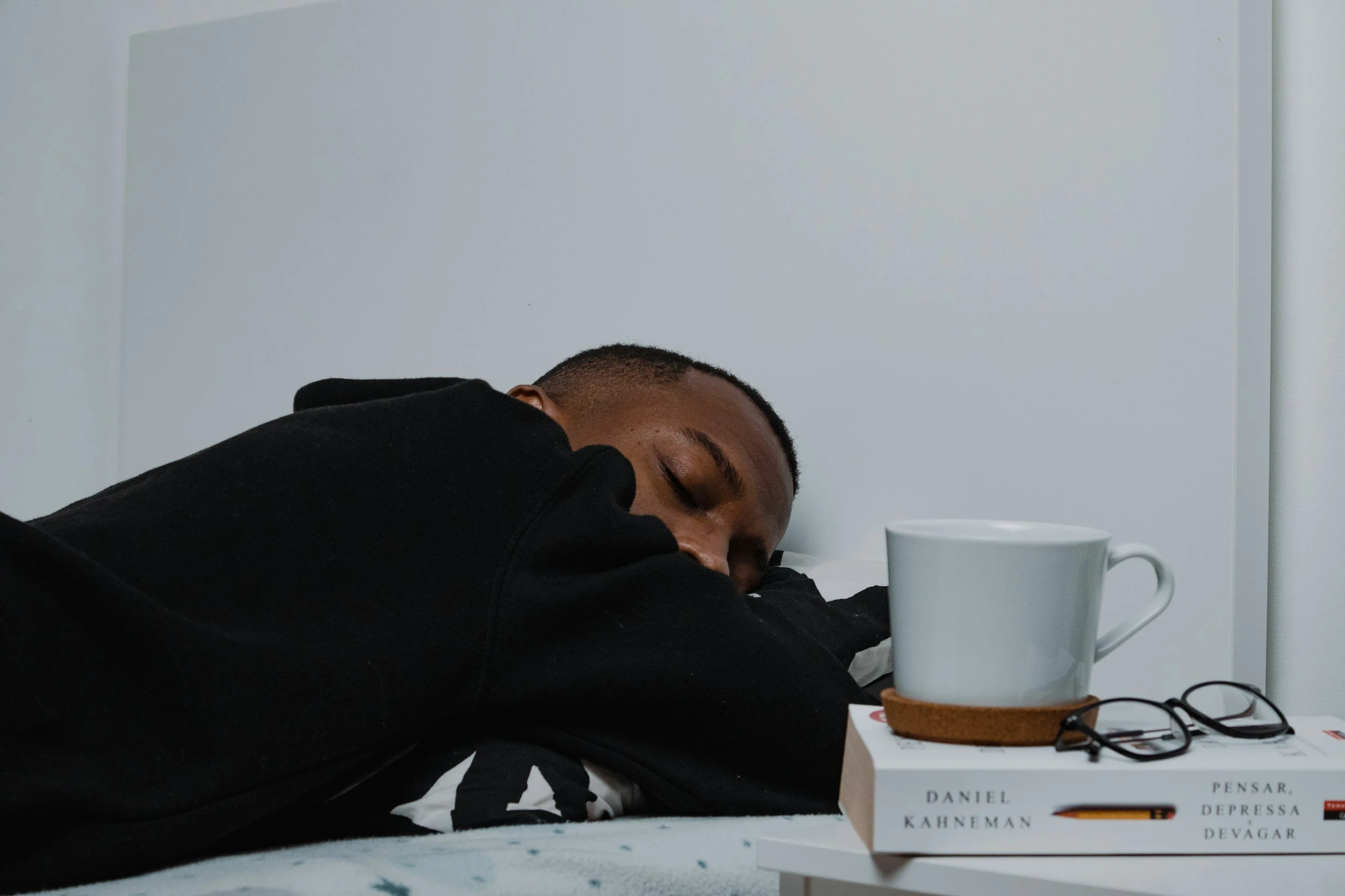A young man of color with short hair sleeping on a white bed with a white wall in the background. There is a white mug, a pair of glasses, and a book on a white nightstand next to him..