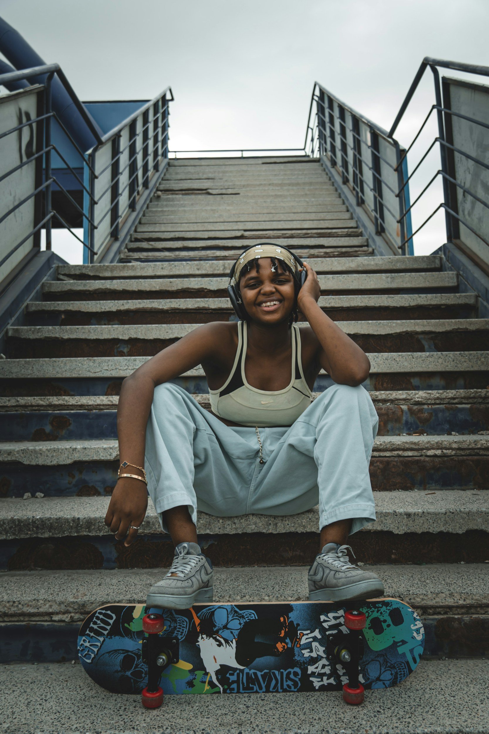 A smiling young person wearing headphones, sitting on outdoor stairs with skateboarding equipment including a skateboard with colorful graphics and red wheels.