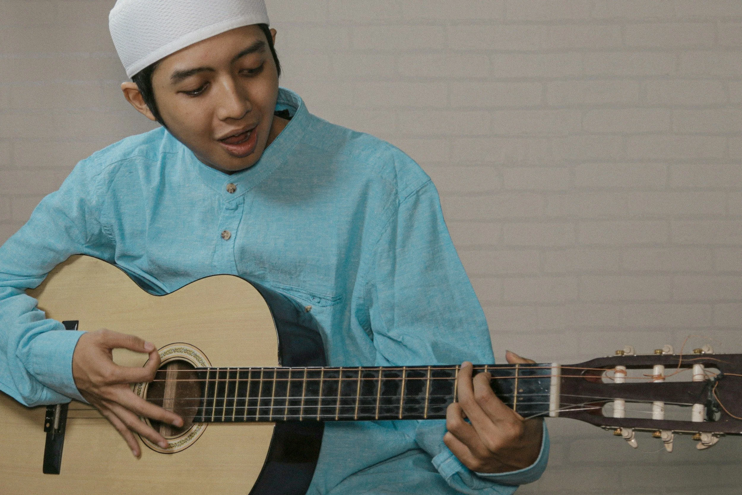 A young man wearing a white cap and blue shirt, playing an acoustic guitar.