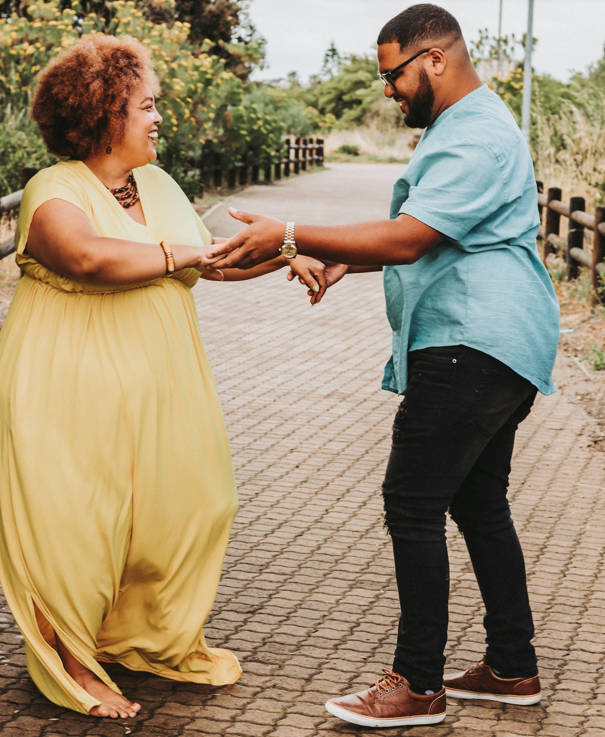 A woman in a yellow dress and a man in a blue shirt are holding hands and smiling at each other outdoors on a brick-paved walkway surrounded by greenery.