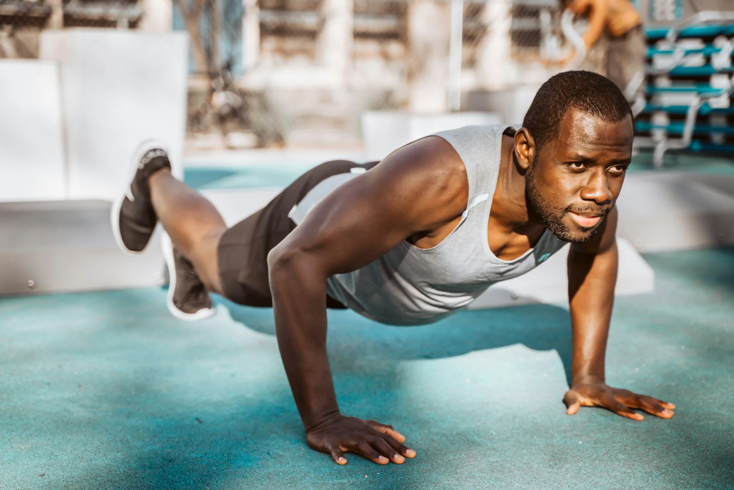 A man in athletic clothing is performing a push-up exercise on a green gym mat indoors, with a blurred background of gym equipment and a pool.