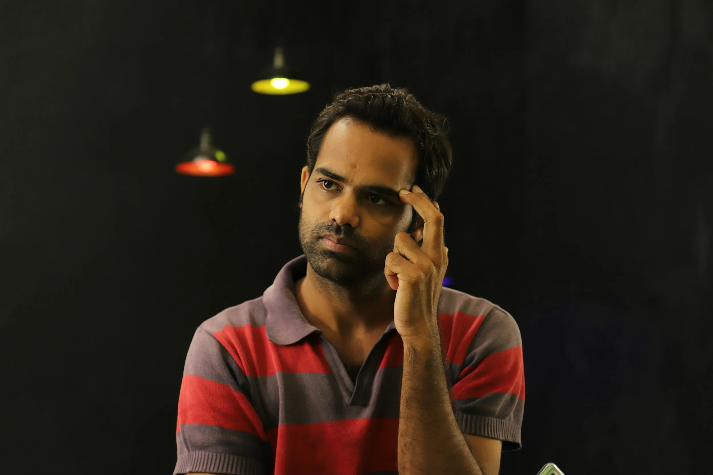 A young man with dark hair and a beard is fixing his glasses while looking to the side against a dark background with colorful ceiling lights.