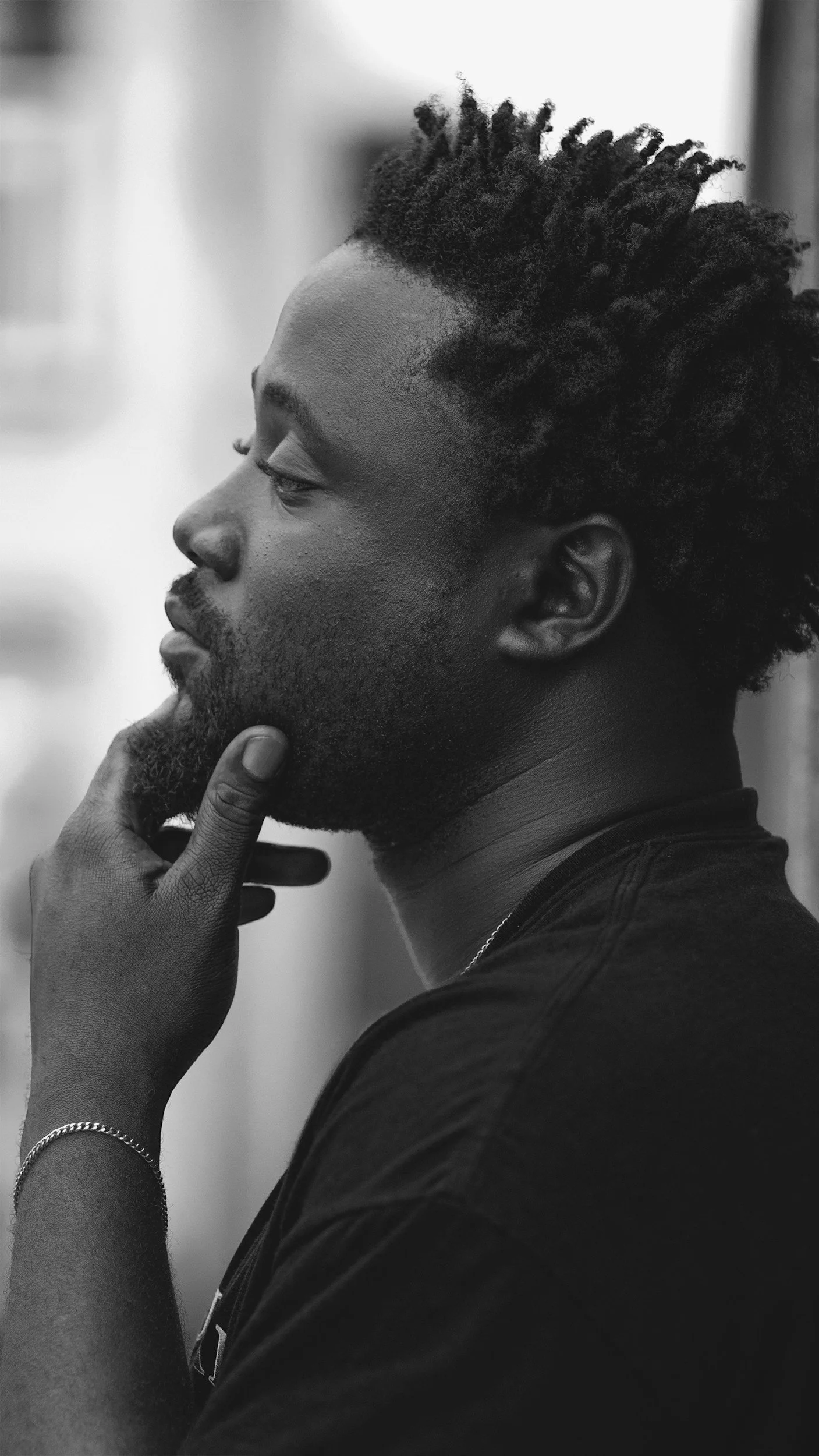 Black and white profile of a man with textured dreadlocks, beard, and mustache, touching his chin with his right hand, wearing a dark shirt and a bracelet on his wrist, against a blurred background.