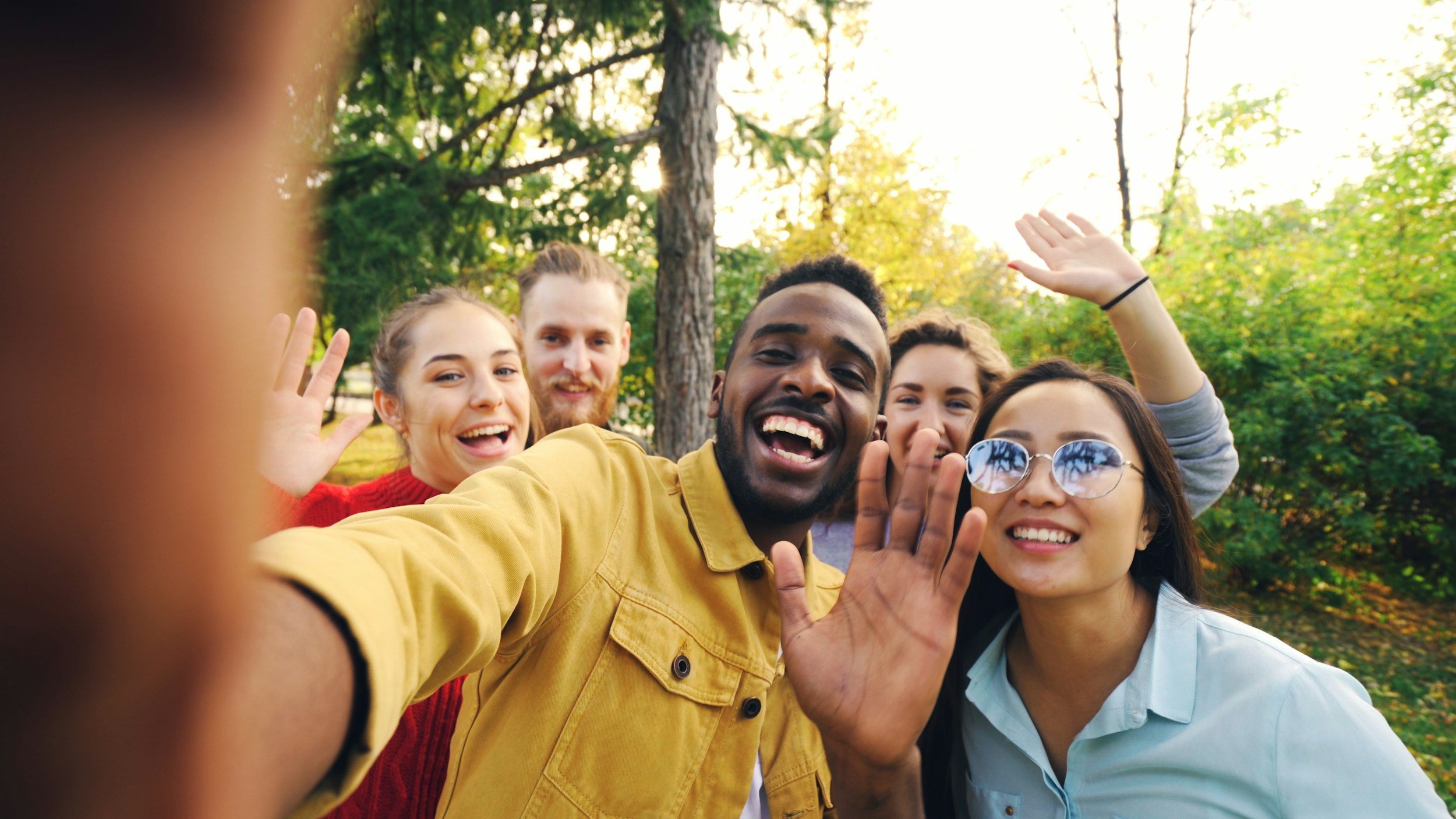 A diverse group of young adults taking a selfie outdoors in a park with autumn trees in the background.