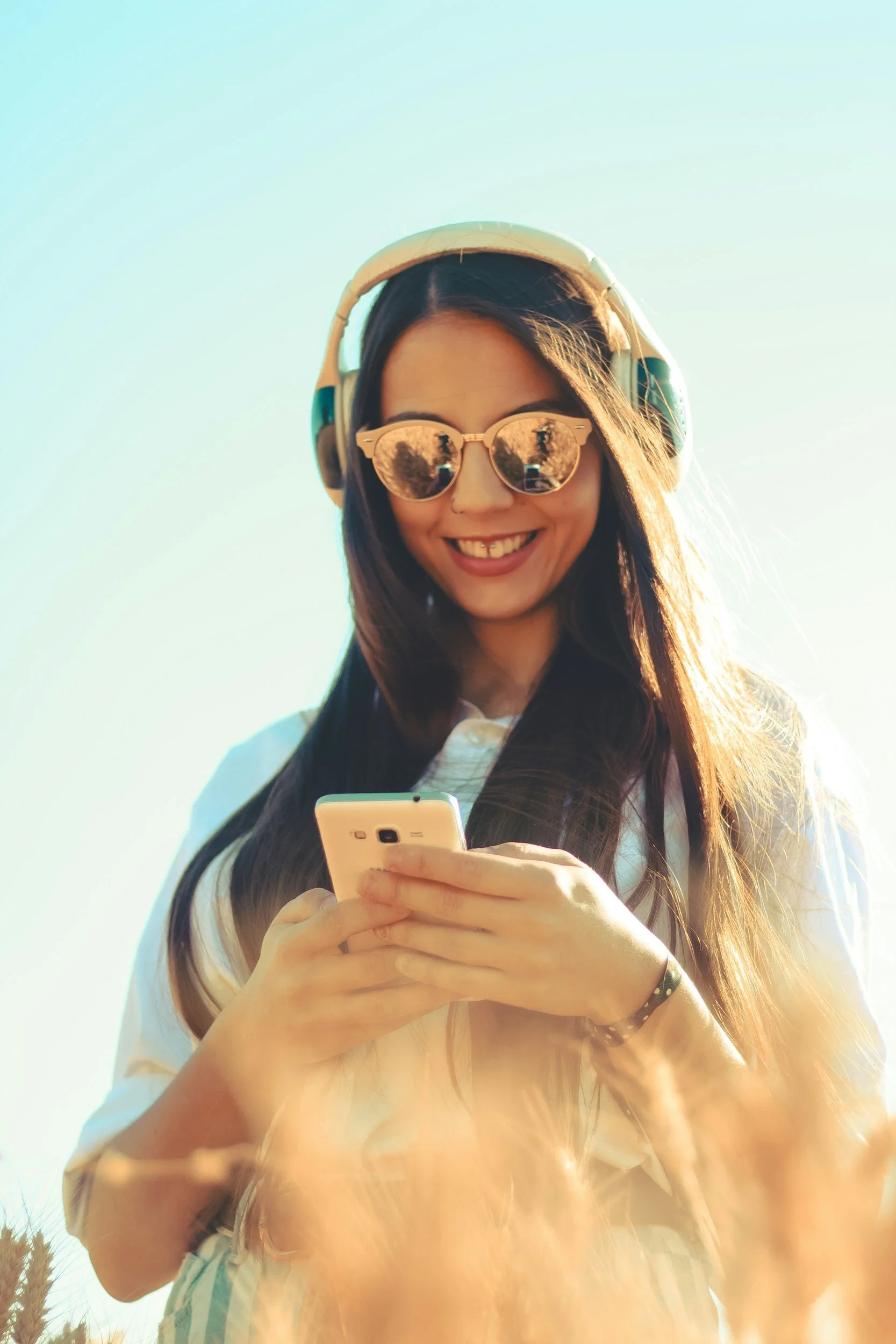 A young woman of color with long dark hair is smiling while looking down at her phone. She is wearing round sunglasses that reflect the outdoor scenery, a wide-brimmed hat, and a light-colored, striped shirt. The background shows a clear blue sky.