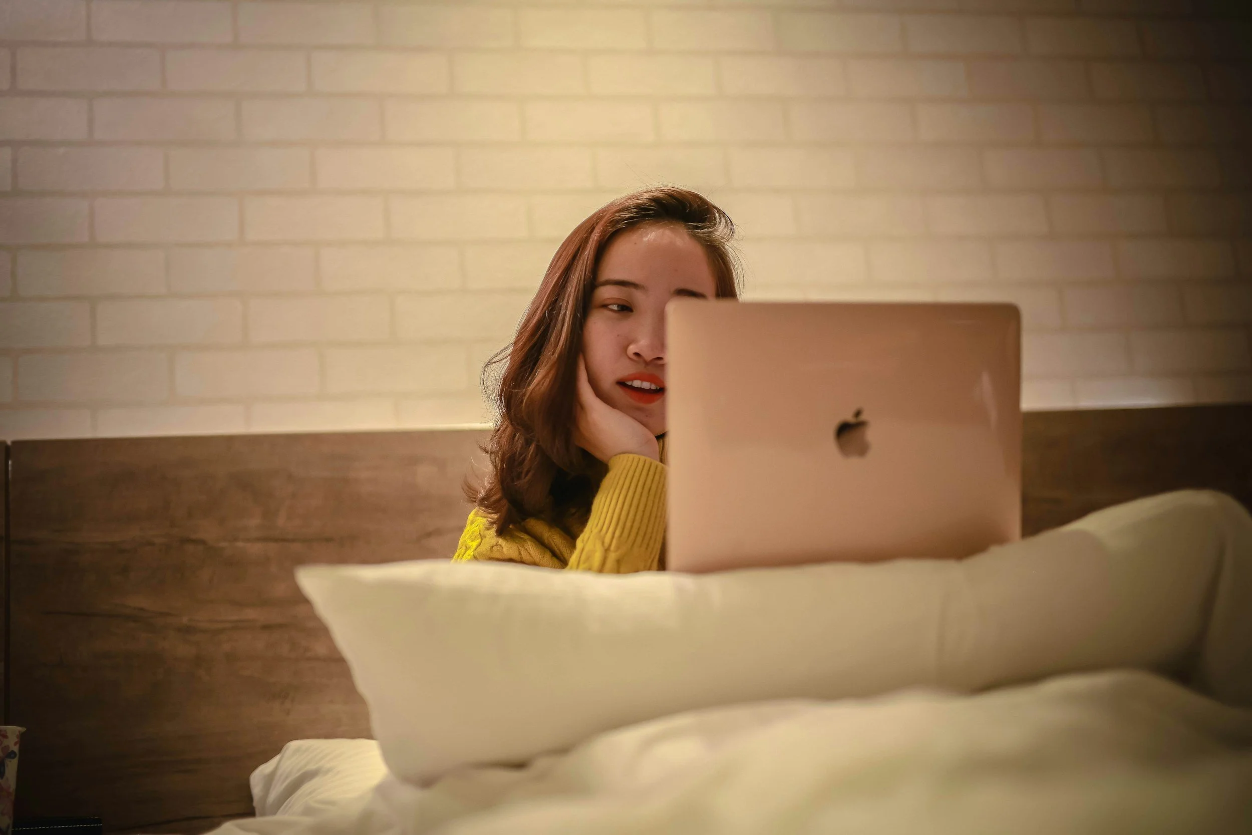 A young woman of color with brown hair, wearing a yellow sweater, looks at her reflection in a mirror in a warmly lit room with light-colored brick walls.