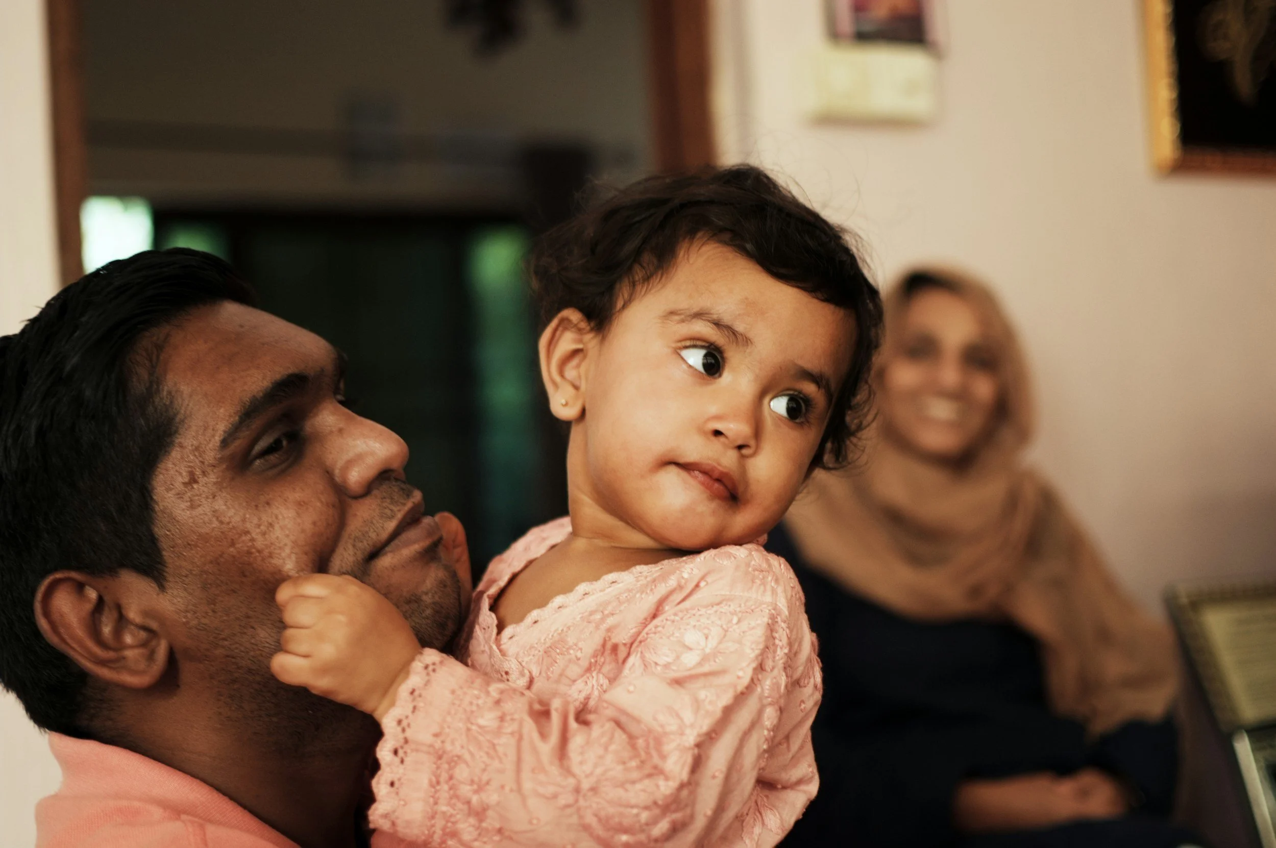 A man holding a young girl with curly hair and brown skin, both looking to the right, with a woman in the background smiling.