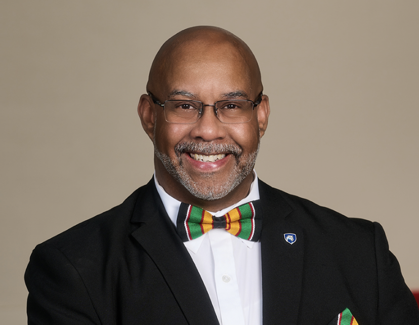 A smiling man of color in a black suit, white shirt, and colorful bow tie, wearing glasses, against a plain background.