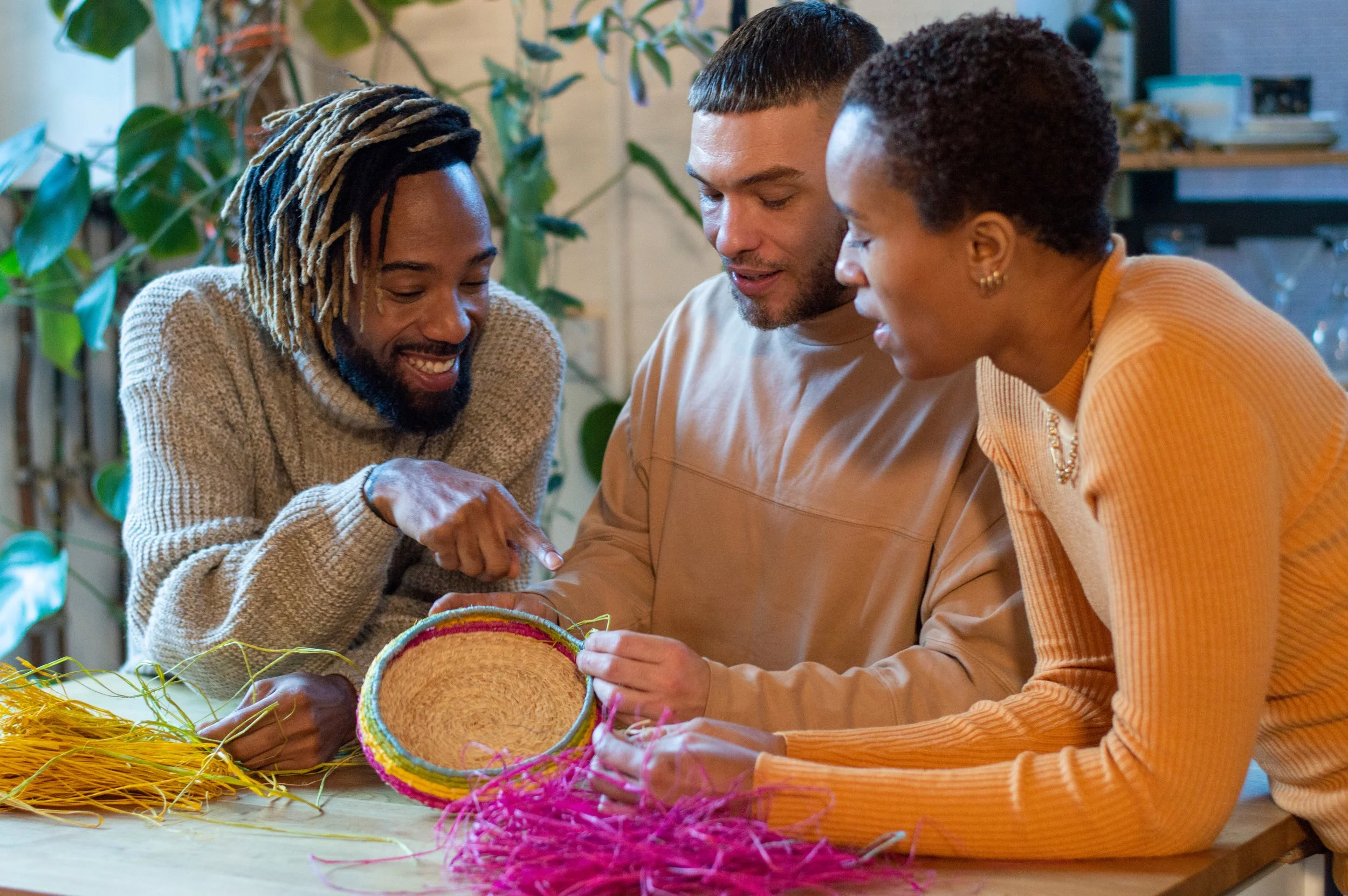 Three people of color are sitting around a table, weaving colorful straw baskets, surrounded by yellow and pink straw and green threads.