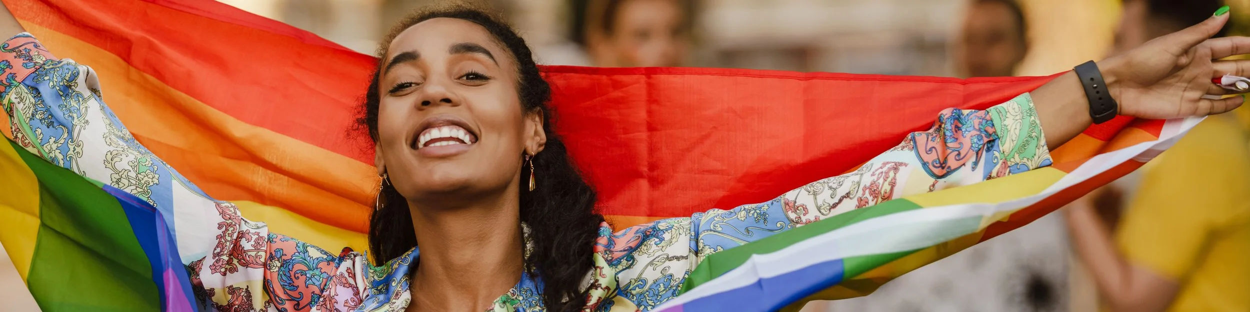 A young woman with curly dark hair smiling and holding a rainbow pride flag during a celebration.