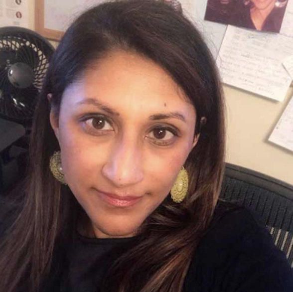 A woman of color with long dark hair and large earrings taking a selfie in an office. Behind her are papers on the wall and a black fan.