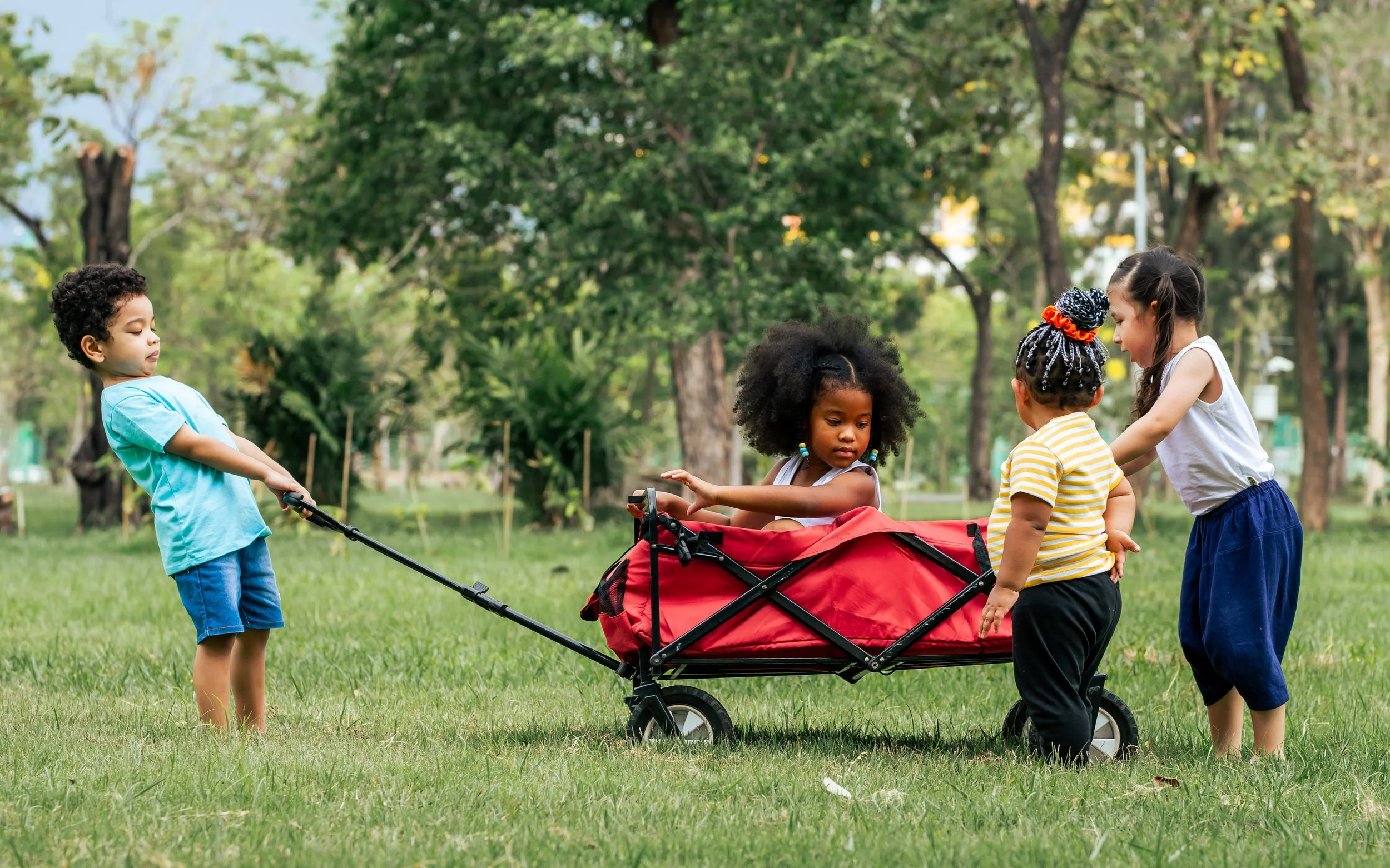 Four young children playing outdoors in a park with trees and grass, two girls sitting in a red stroller, one boy pulling it, and another girl standing nearby.