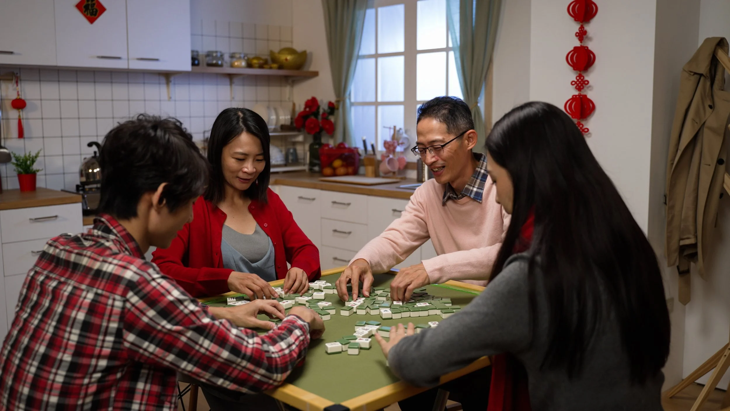 Group of five people playing Mahjong at a table in a kitchen decorated with red Chinese New Year decorations.