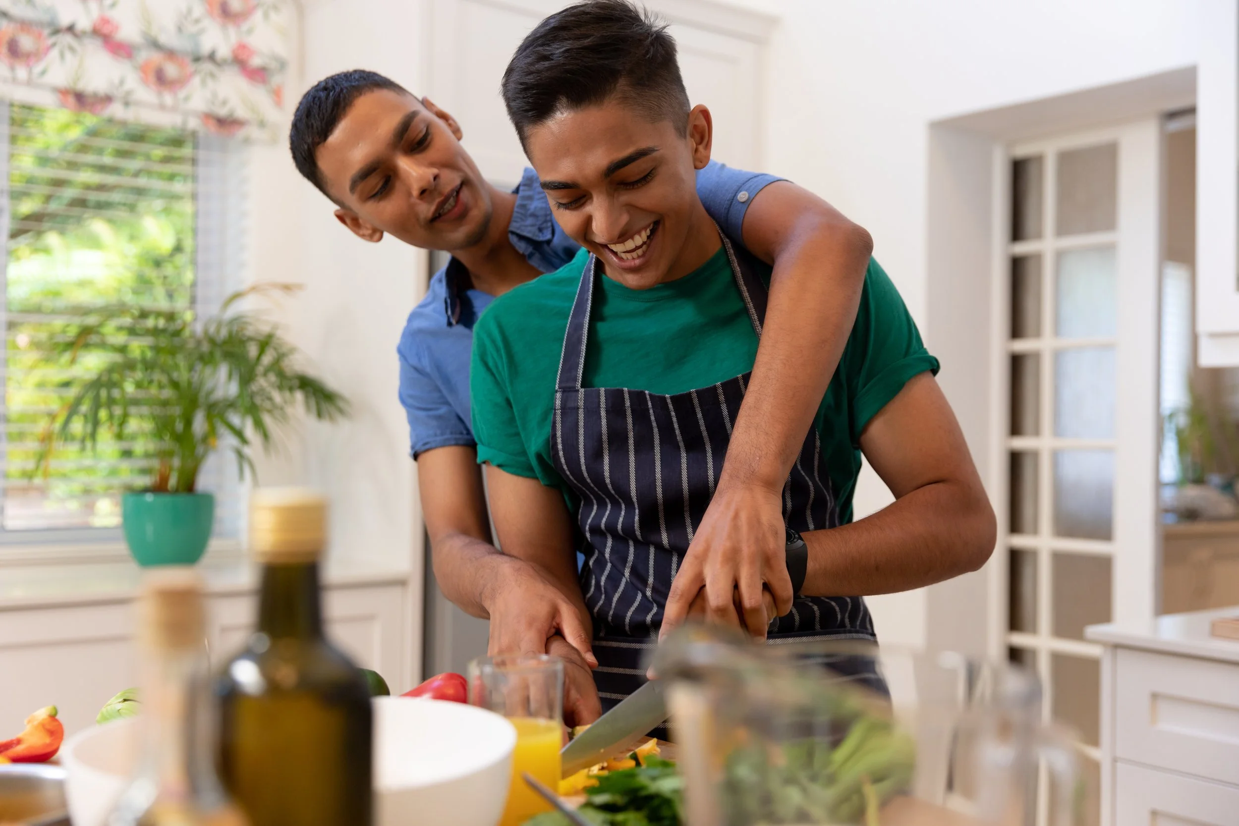 Two men of color in a kitchen are preparing food, one wearing an apron, smiling and working together.