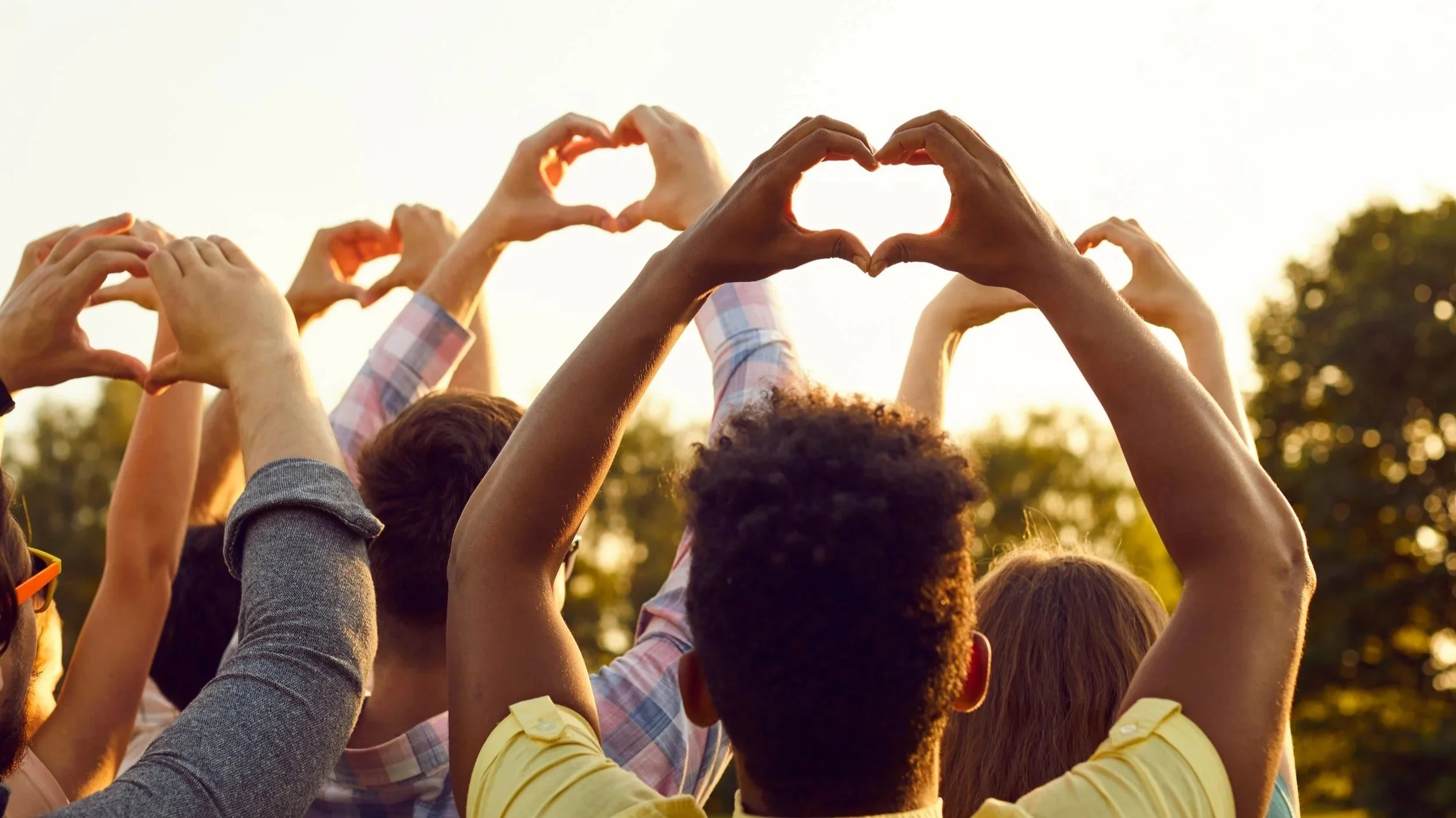 A diverse group of people outdoors making heart shapes with their hands during sunset.