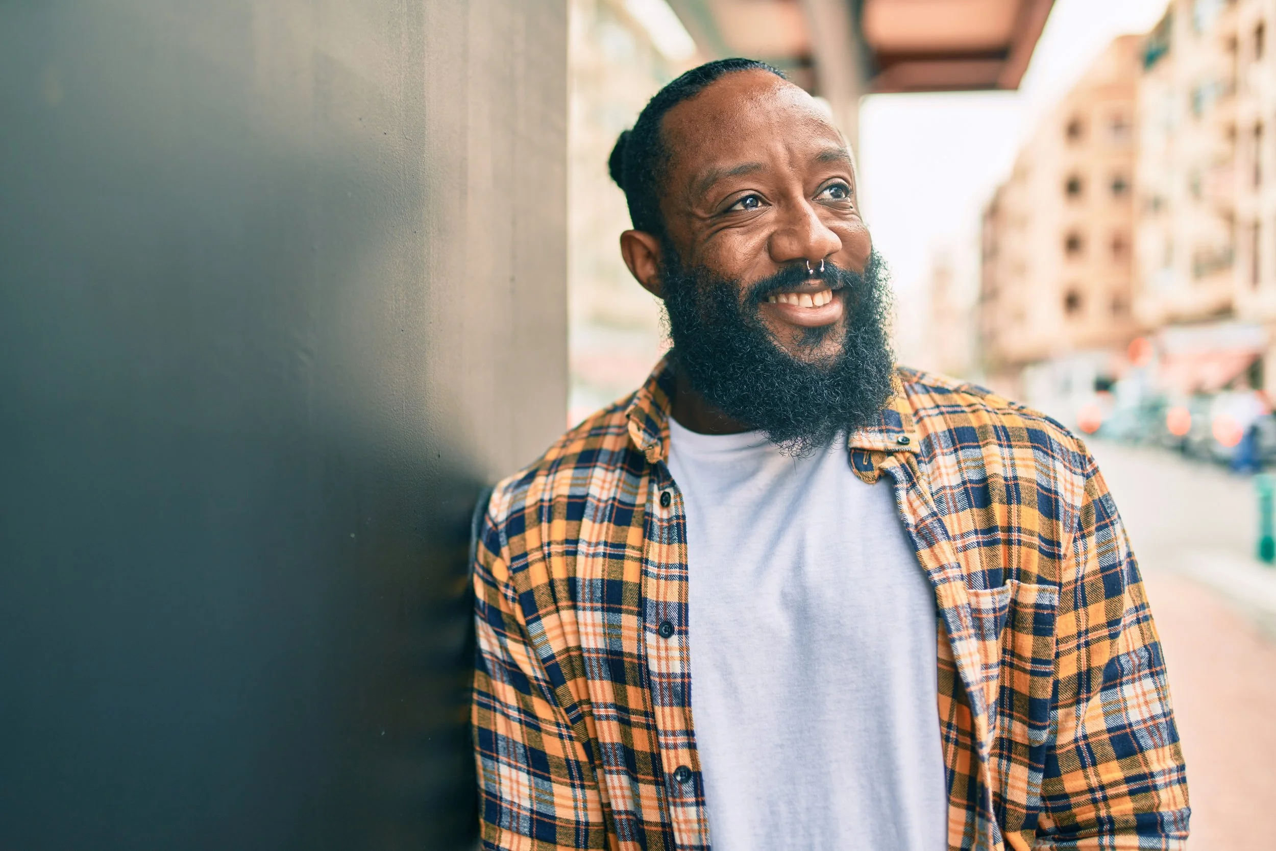 A happy man of color with a beard and a septum piercing is standing outdoors, smiling and leaning against a wall, with a cityscape in the background.
