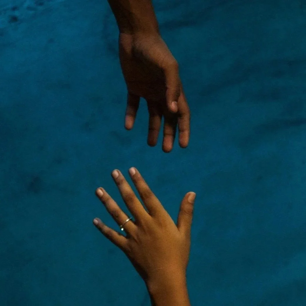 A close-up of two brown-skin hands reaching towards each other against a blue background, with one hand above and the other below.