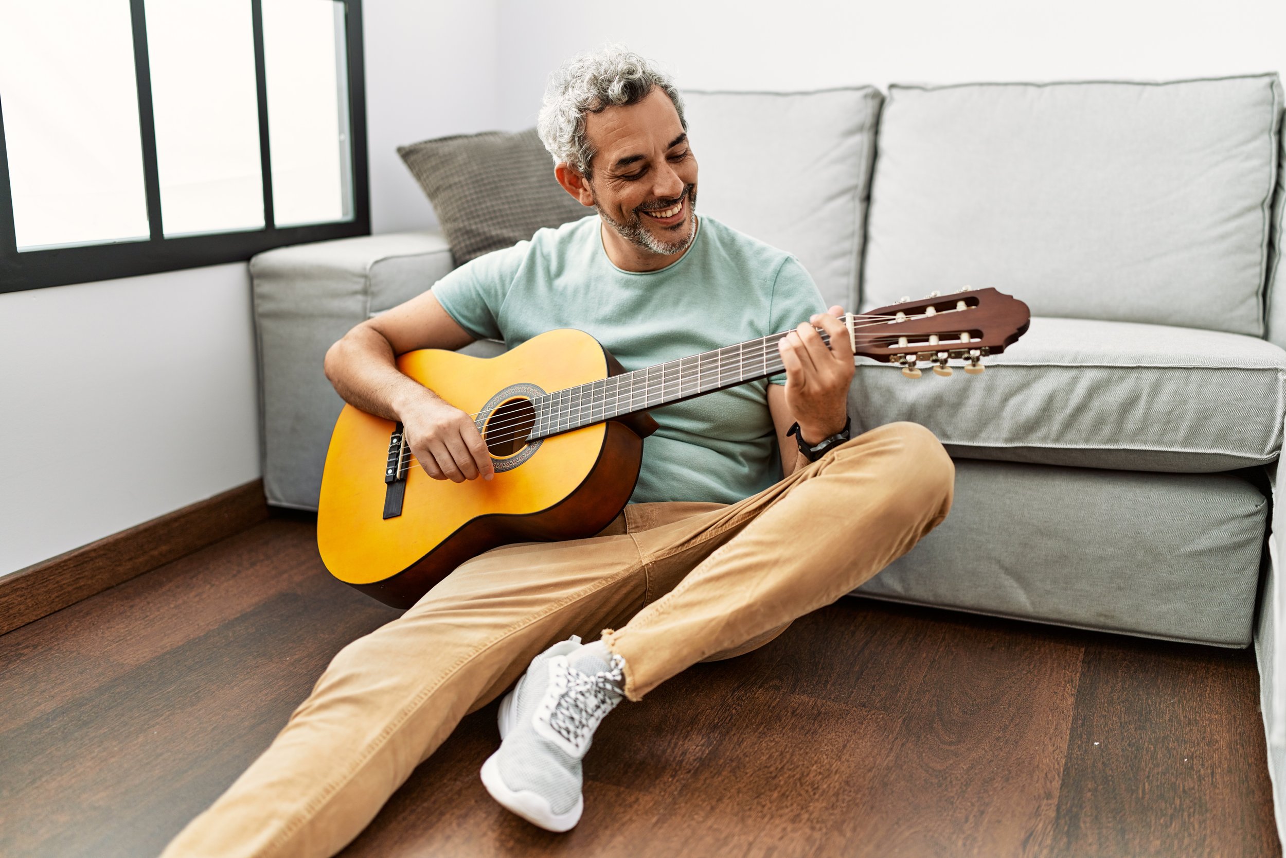 Middle-aged man of color sitting on the floor, playing an acoustic guitar, smiling, in a bright, modern living room with a gray sofa, a gray pillow, and a large window.