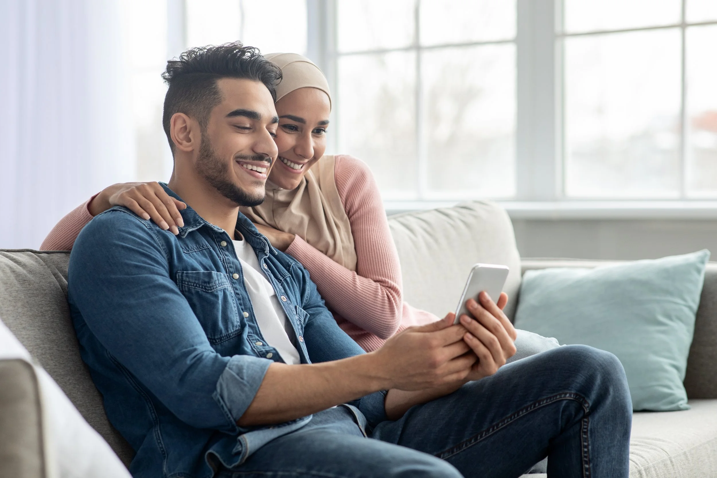 A young man and woman of color sitting on a beige couch, looking at a smartphone together, smiling, in a bright room with large windows.