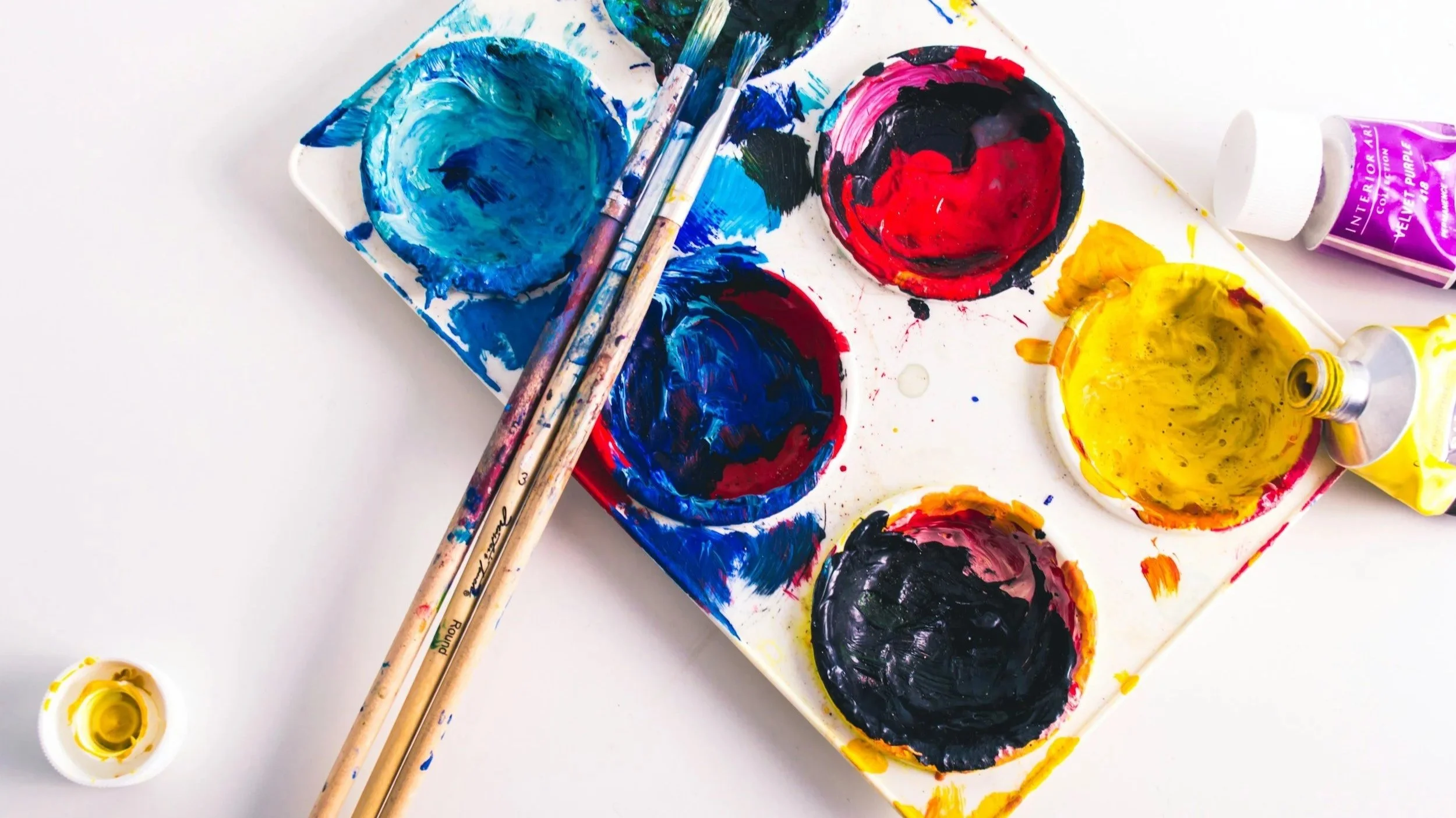 Paint palette with various colors including blue, red, yellow, and black, with paintbrushes resting on the palette and small containers of yellow paint nearby.