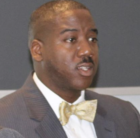 A man of color with short hair wearing a dark suit, white shirt, and gold bow tie.
