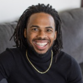 A smiling man of color with dreadlocks, wearing a black turtleneck and a gold chain, sitting on a gray couch.