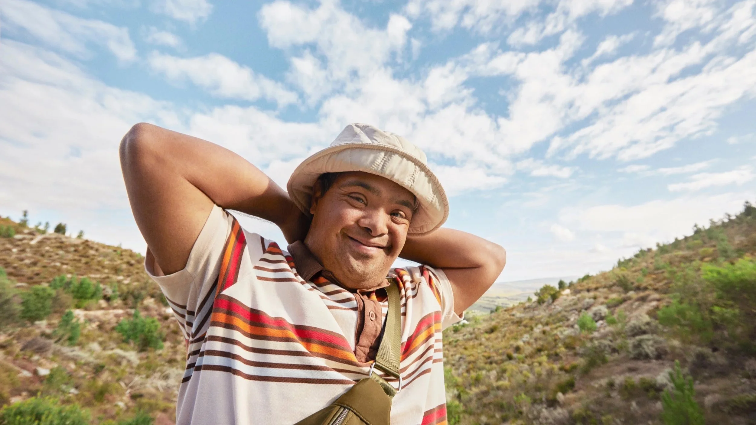 A man of color smiling with arms behind his head, wearing a striped shirt and a hat, standing outdoors with mountains and a cloudy sky in the background.