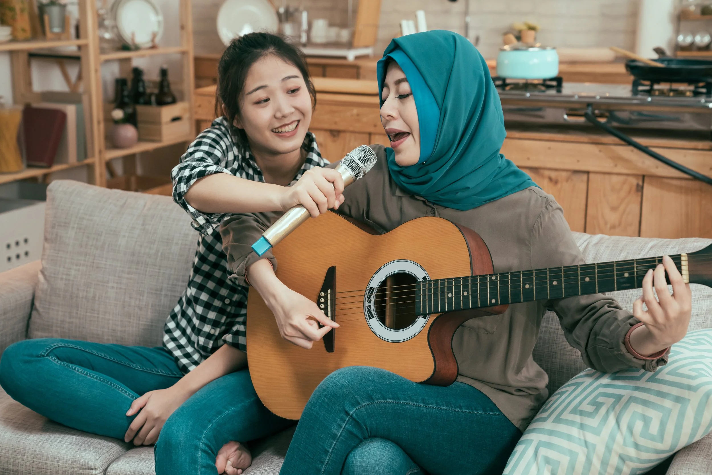 A young woman in a checkered shirt and another woman wearing a blue hijab sit on a couch, singing and playing guitar together in a cozy, wooden home setting.