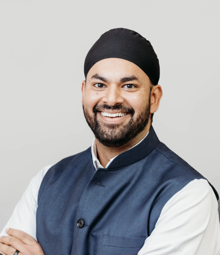 A smiling man of color wearing a black turban, white shirt, and blue vest, standing against a light gray background.