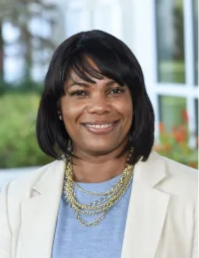 Professional woman of color with short black hair smiling, wearing a cream blazer and layered gold necklaces, in a bright indoor setting with windows and greenery in the background.