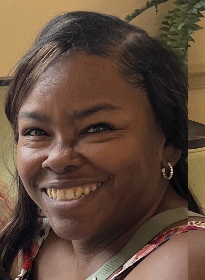 Close-up of a smiling woman with dark hair, wearing earrings, and a floral top, indoors with a plant in the background.