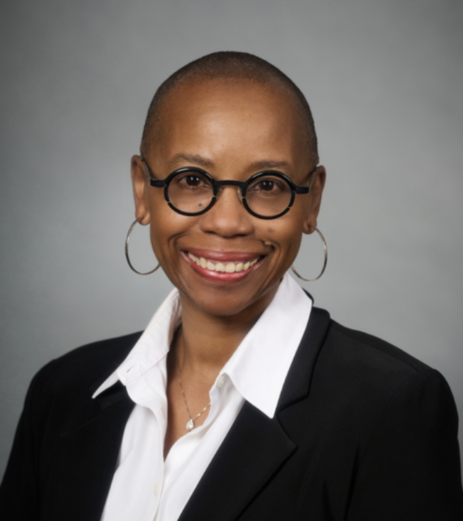 Smiling woman of color with glasses, hoop earrings, short hair, white shirt, black blazer, and a necklace against a gray background.