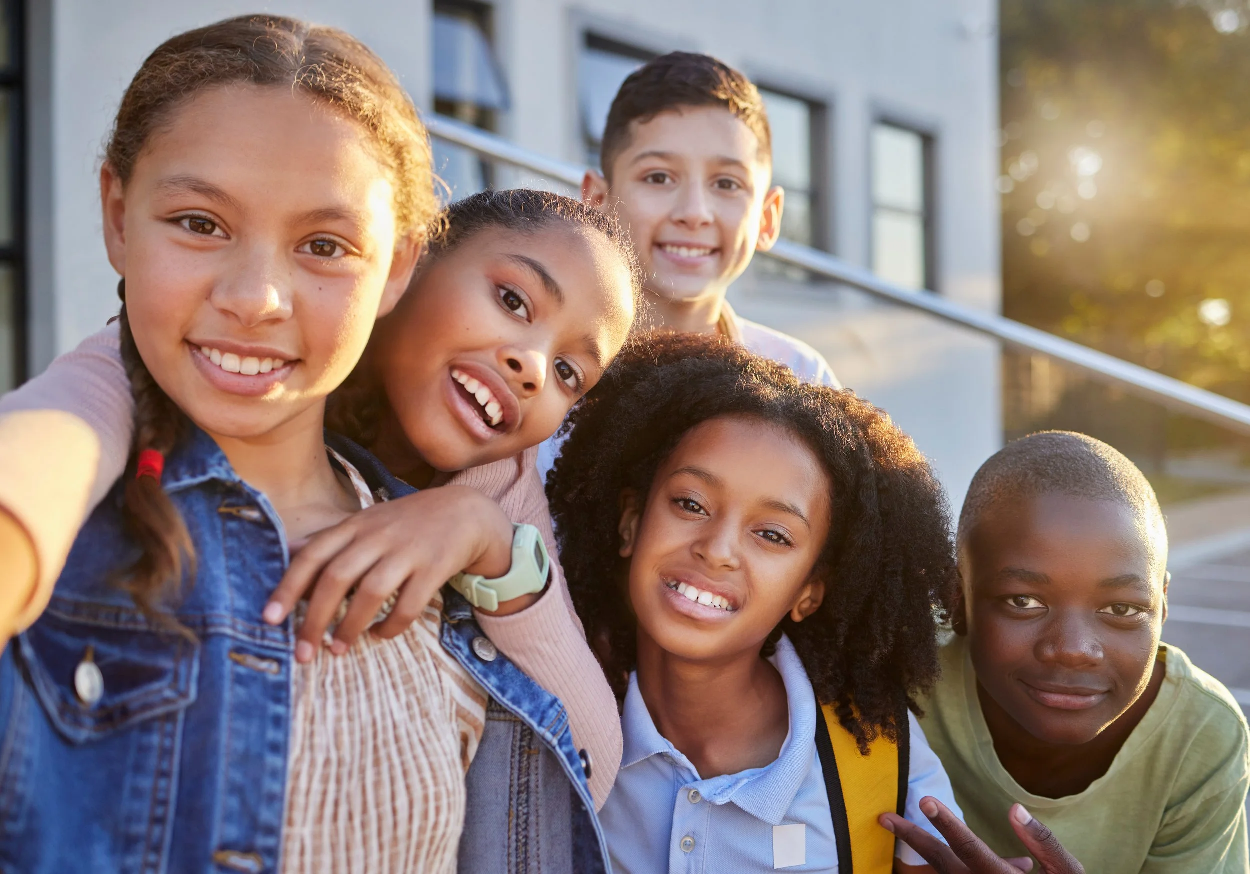 A group of diverse children smiling and taking a selfie outdoors in the late afternoon or early evening.