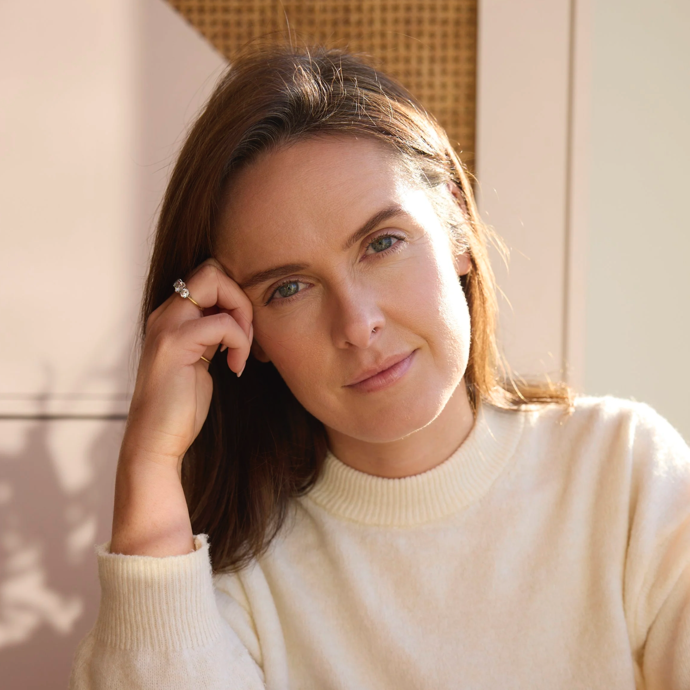 A portrait of Claire Cullen: A woman with brown hair and light eyes, wearing a cream-colored sweater, resting her head on her hand, looking at the camera with a slight smile.