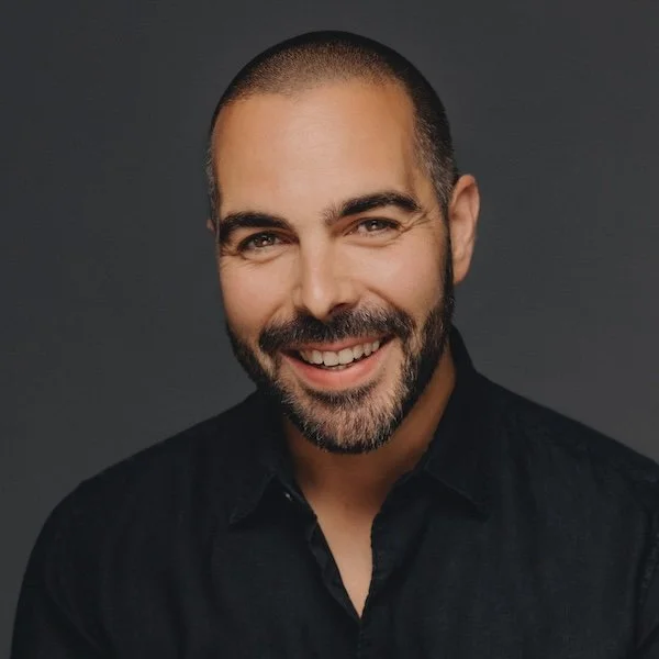 A smiling man with a beard and short hair, wearing a black shirt, against a dark background.