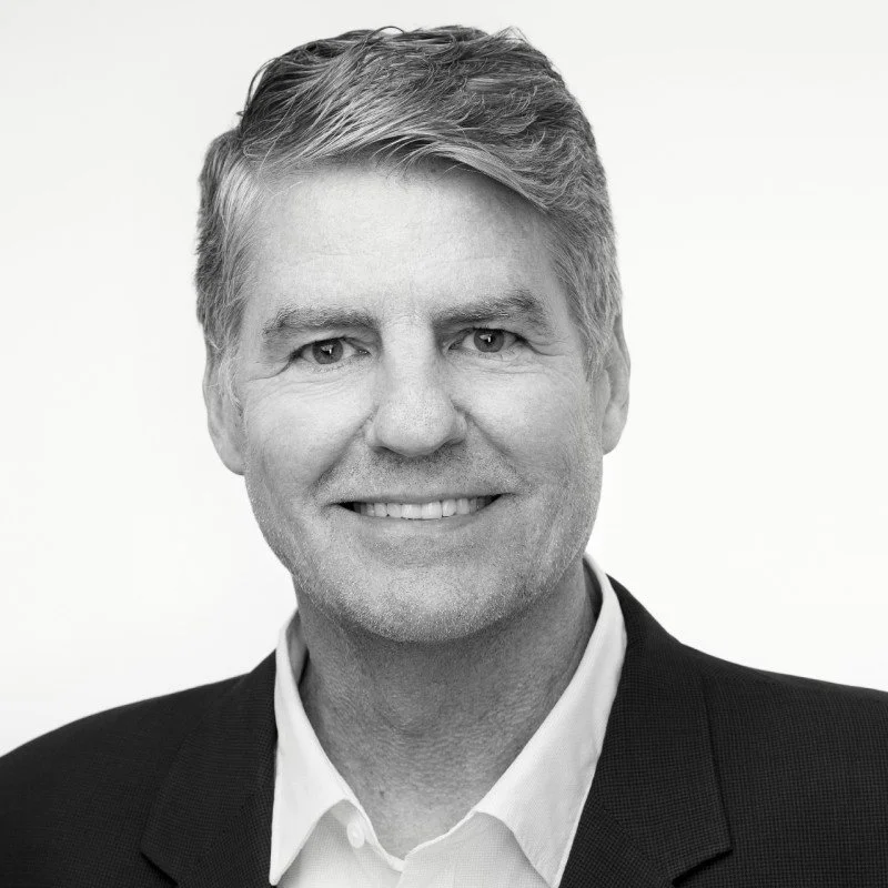 A portrait of Gene Sheridan: A man with short, styled hair, smiling, dressed in a suit and collared shirt.
