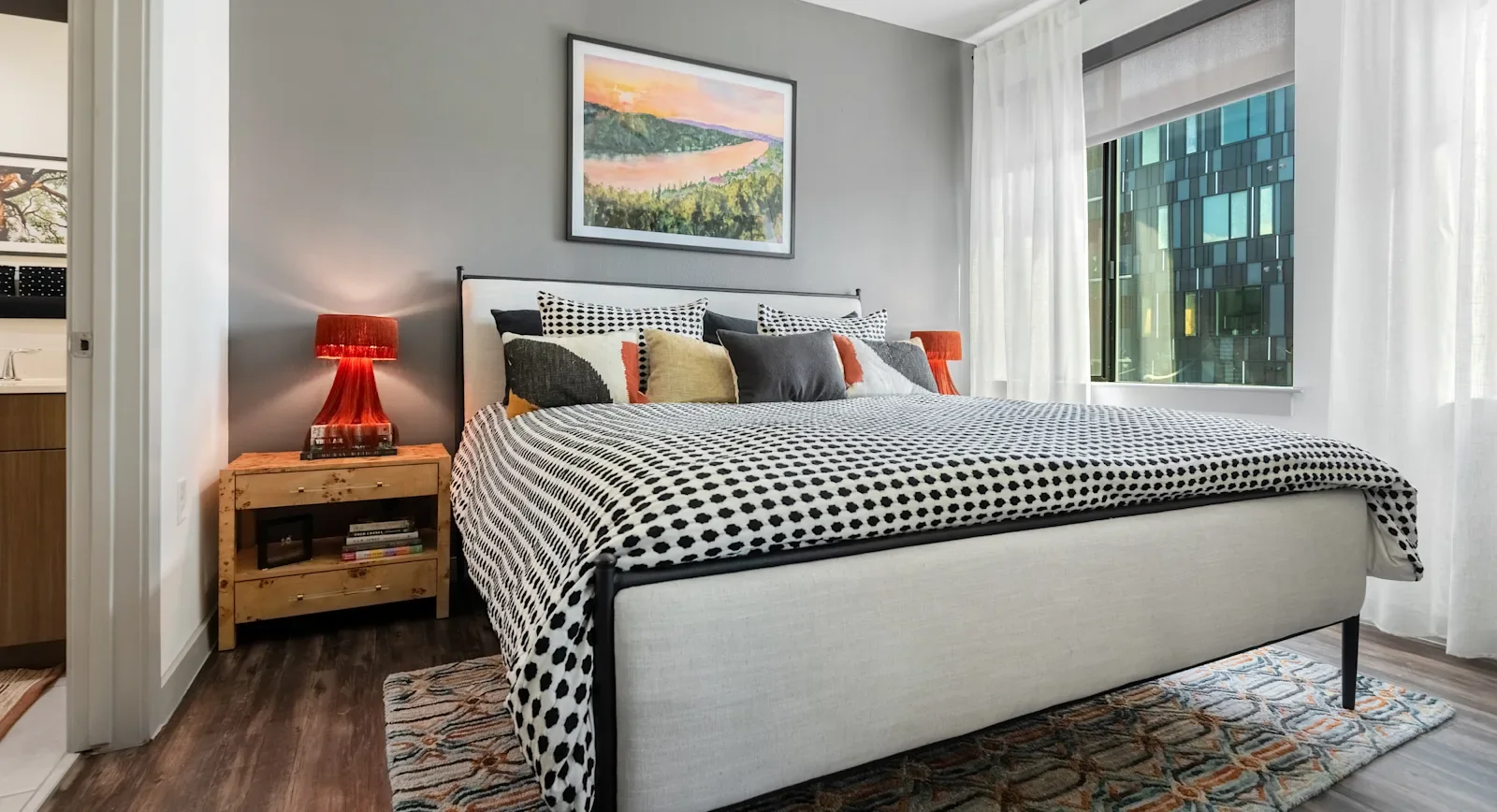 Lamar Union Apartment Bedroom With Polka Dot Bedspread, Red Lamp, Large Window, Landscape Painting, and Colorful Rug