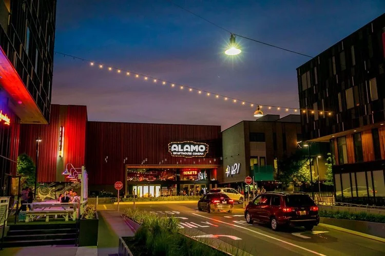 People walking toward the entrance of Alamo Drafthouse Cinema at night with neon signage and outdoor seating