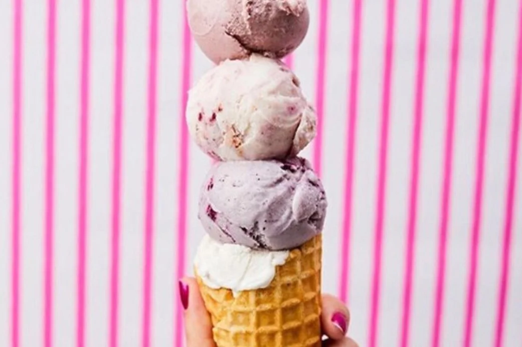 Stack of four colorful ice cream scoops on a waffle cone held against a pink striped background