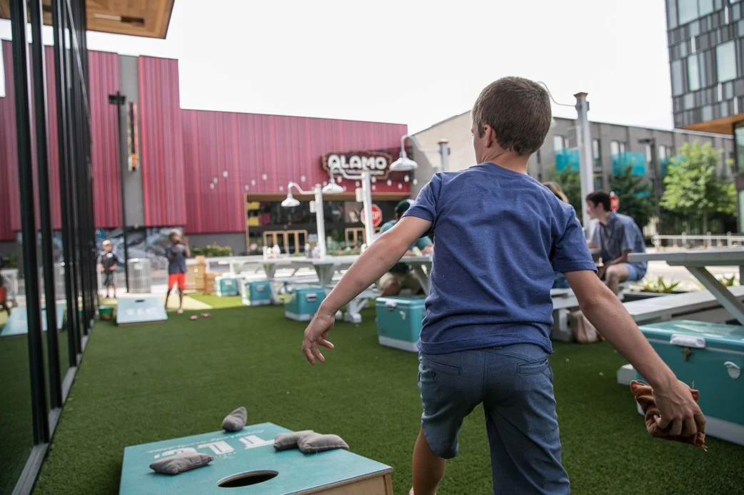 Child playing outdoor cornhole at TLC Lamar Union with Alamo Drafthouse in the background