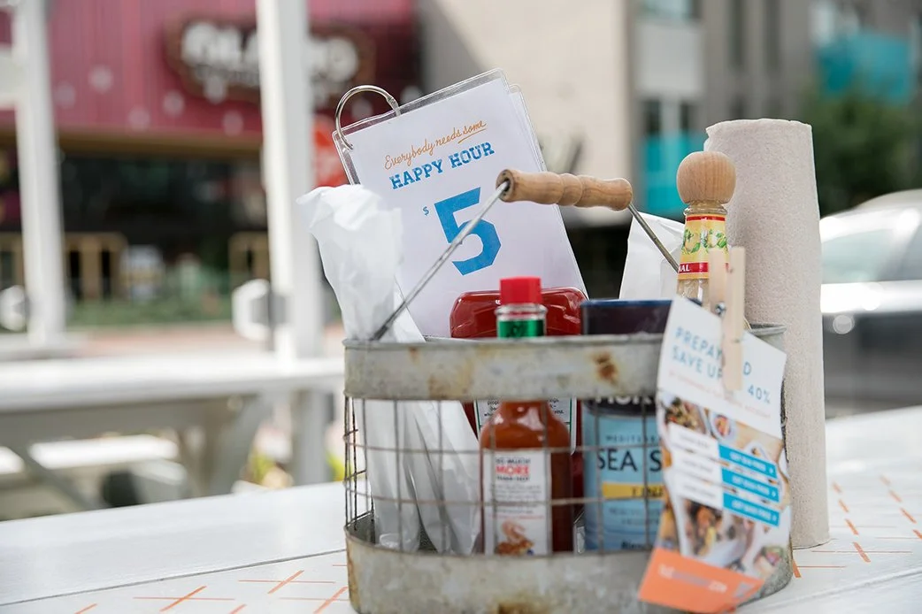 Outdoor table setup with condiments and happy hour sign at TLC Lamar Union