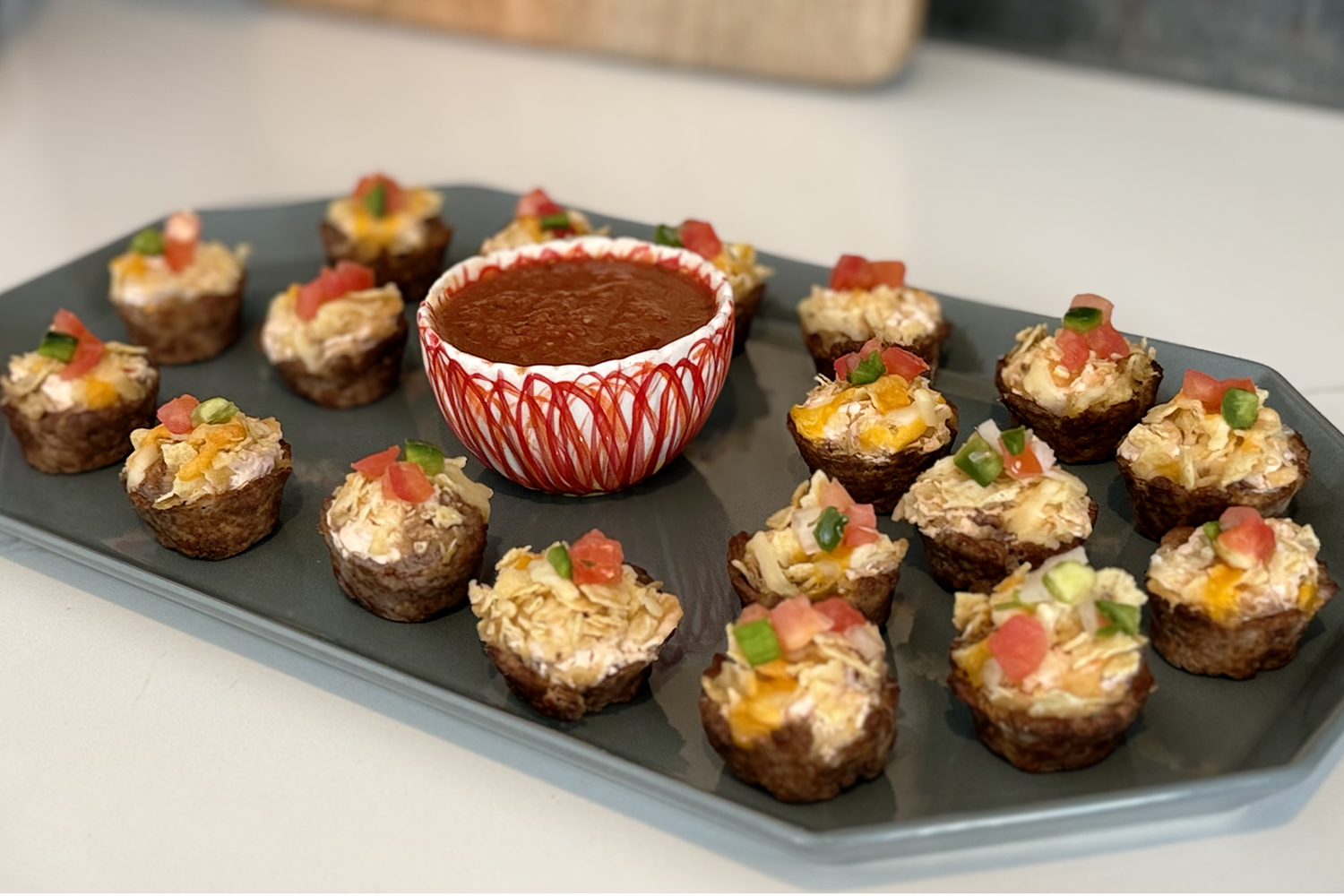 A grey octagonal tray serves mini taco tartlets with salsa at the ready. Topped with beautiful green and red pico de gallo and crushed corn tortillas, the beefy, cheese tart recipe is a tasty addition to your gathering or family meal.
