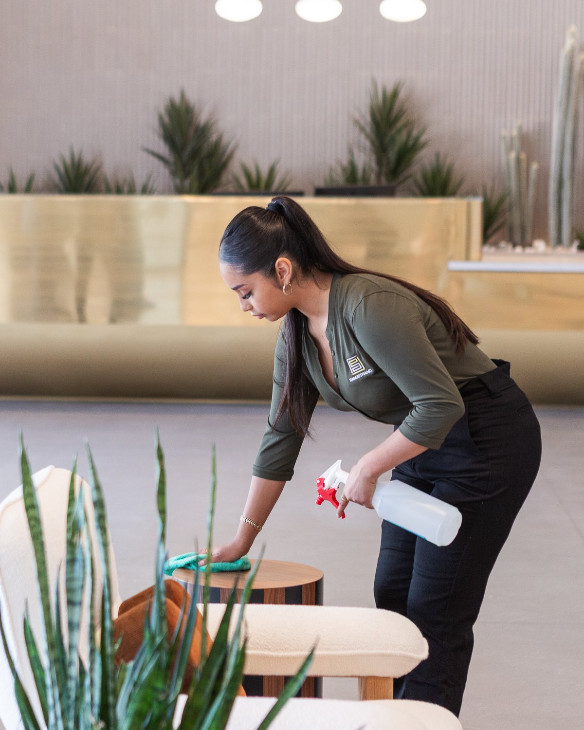Woman cleaning a small round wooden table in a modern, well-lit indoor space.