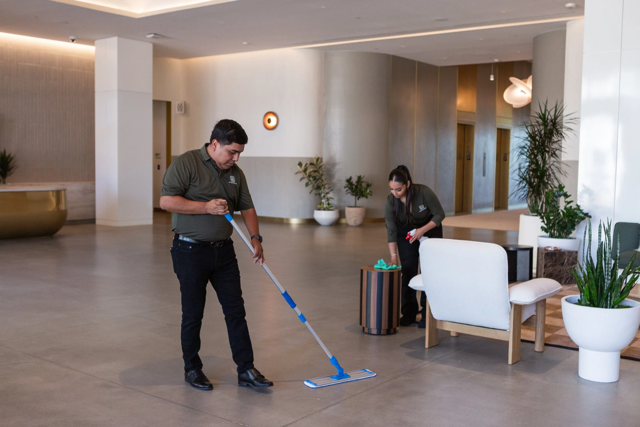Two hotel staff members cleaning the lobby. One man is mopping the floor while a woman is wiping a small table.