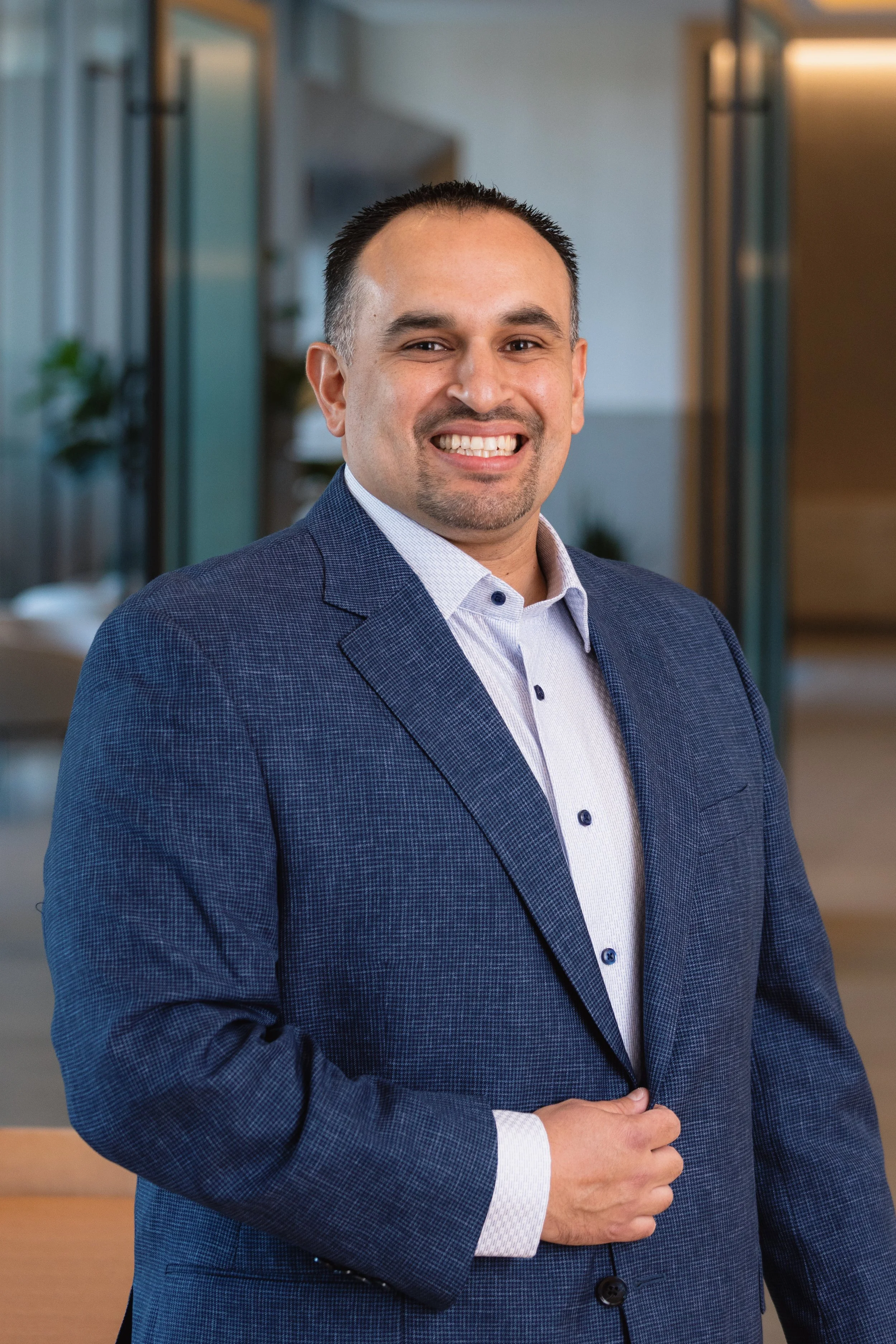A smiling man in a blue suit and light-colored shirt, standing in a modern office setting with glass walls and indoor plants.
