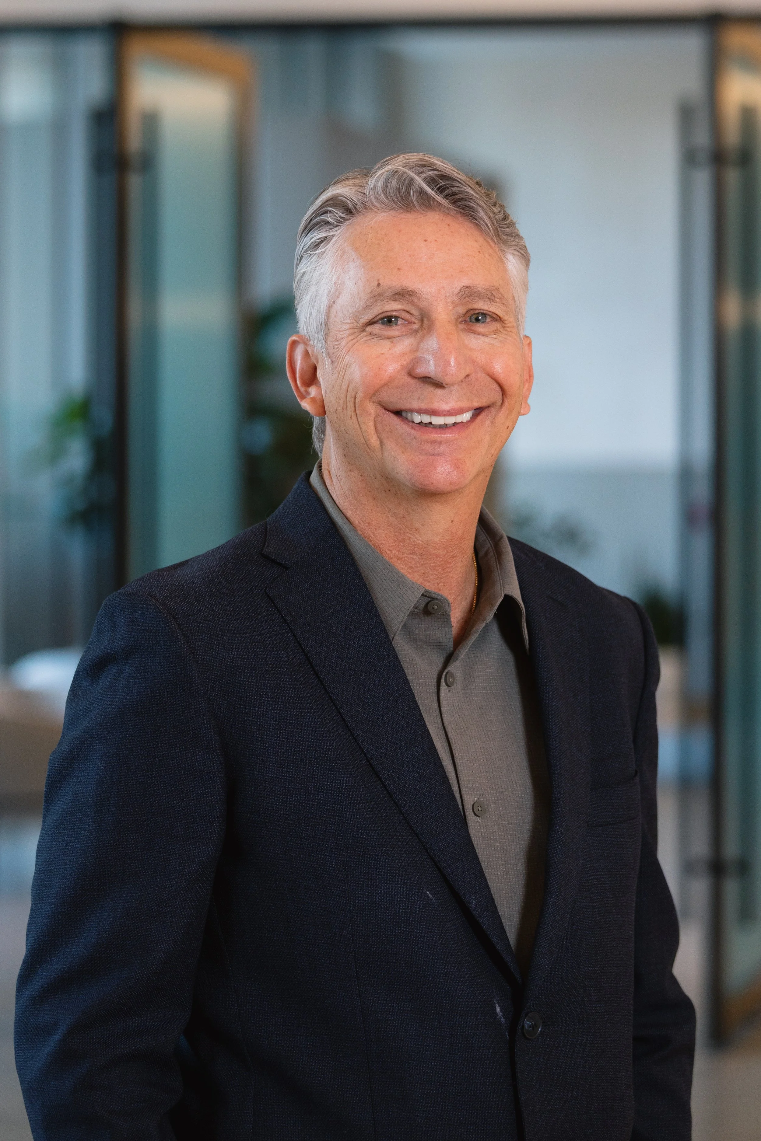 A smiling man in a dark blazer and gray shirt standing in a modern office space.