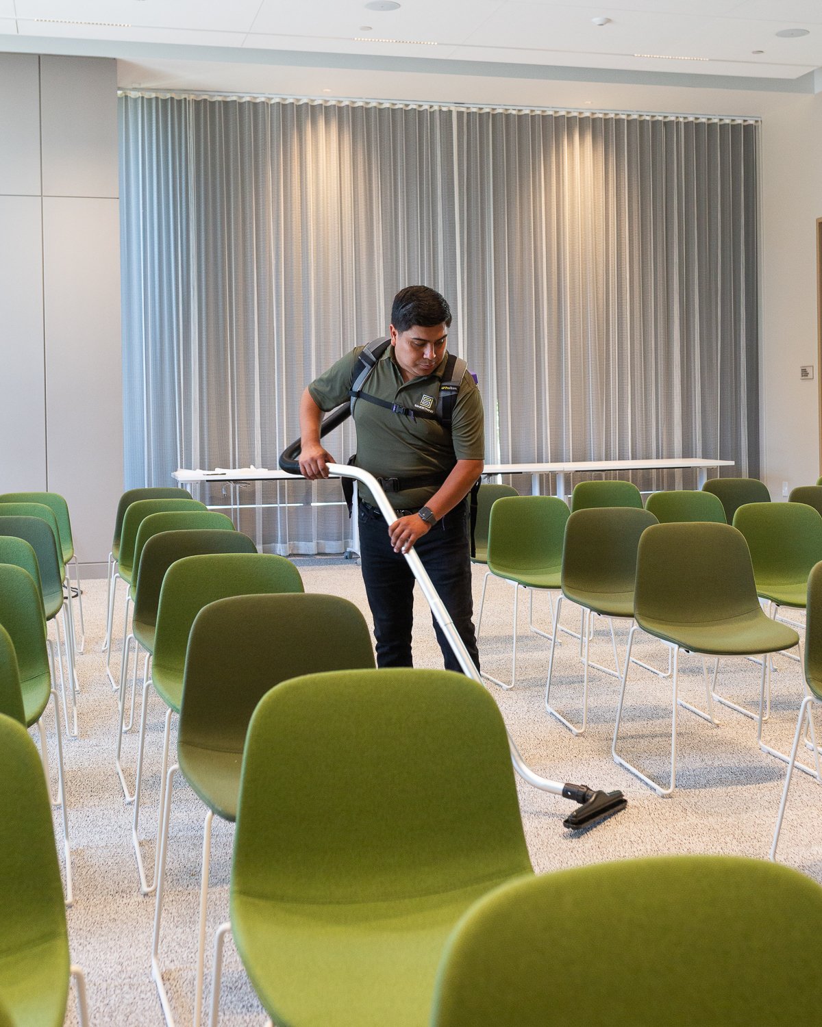 A man cleaning a conference room with green chairs and a gray curtain in the background.