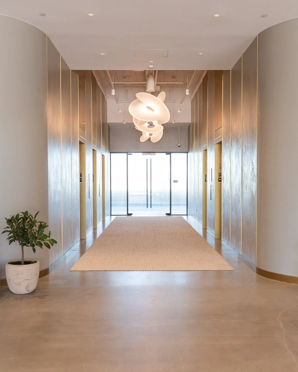 Modern hotel lobby corridor with elevators, a hanging abstract light fixture, a potted plant, and sliding glass doors at the end.