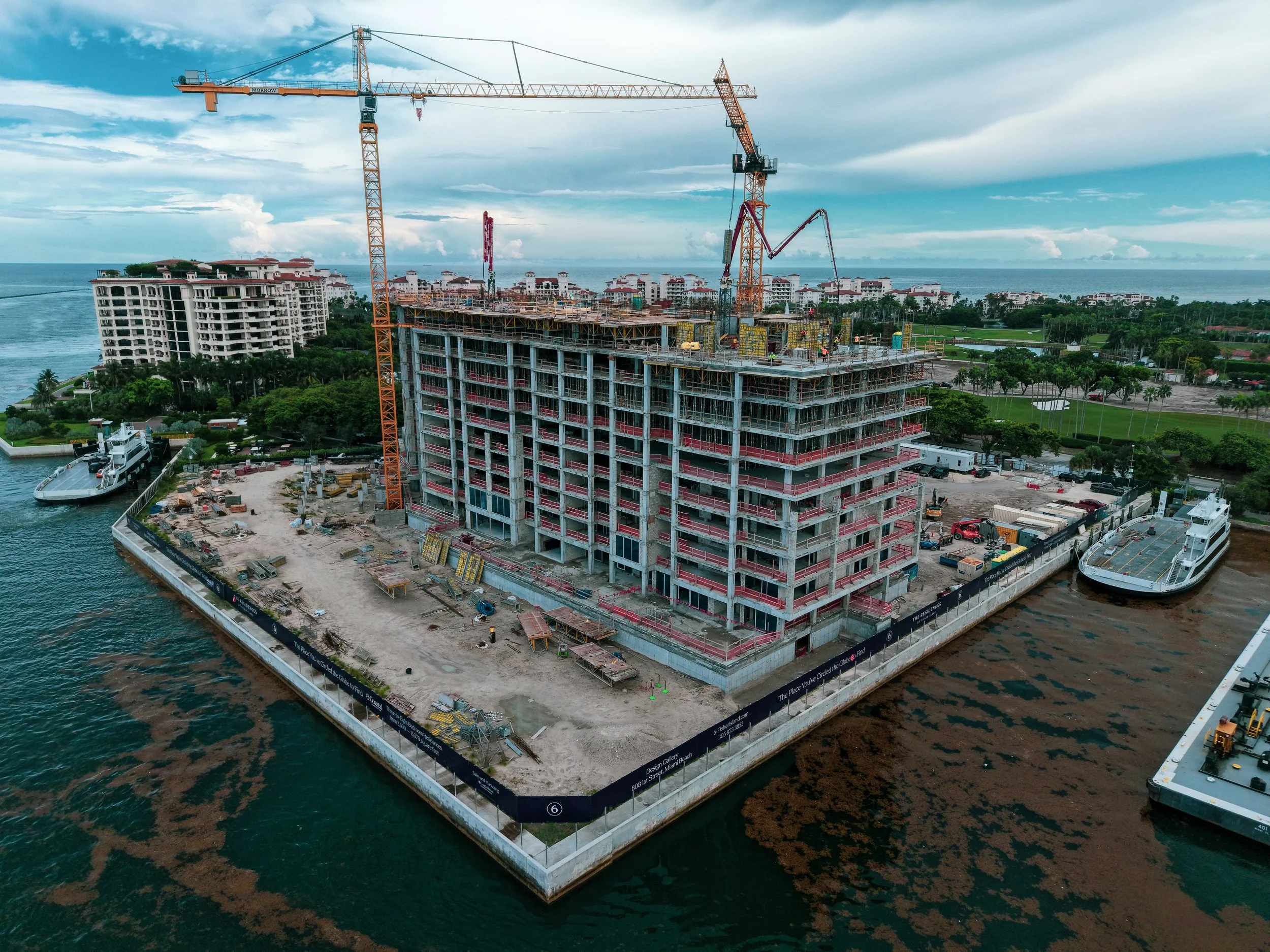 A large construction site on a waterfront, featuring a high-rise building under construction with cranes, scaffolding, and construction equipment, surrounded by palm trees and nearby residential buildings.